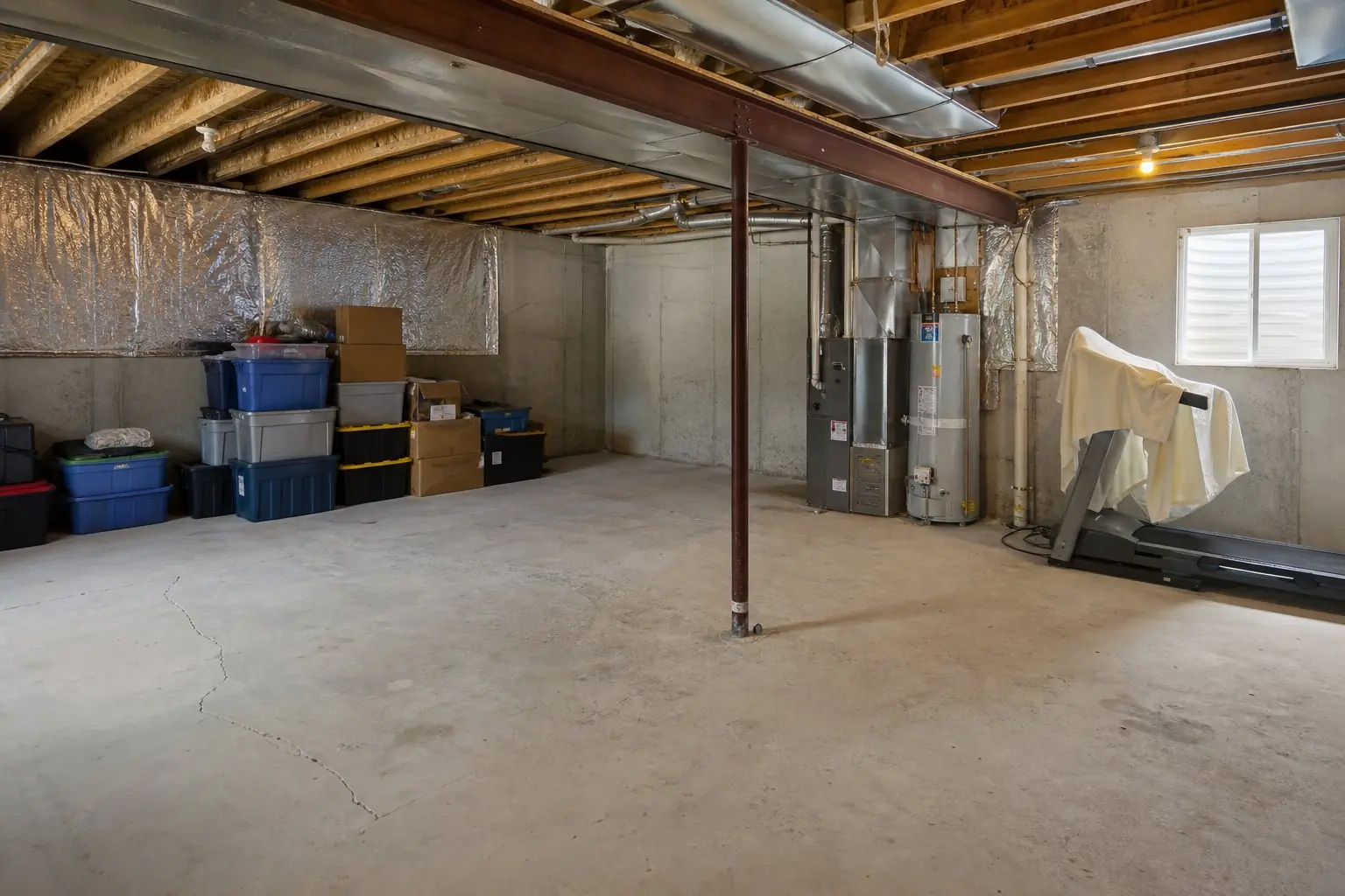 Unfinished basement with stacked storage bins, a steel support column, exposed ductwork, a furnace and water heater, and a treadmill under a sheet